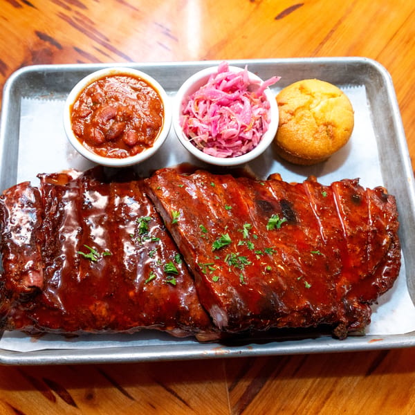 Big Daddy's NY BBQ ribs with baked beans, pink coleslaw, and cornbread on a metal tray.